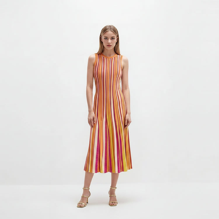 A woman stands against a plain white studio background wearing a sleeveless midi dress with vertical pink, orange, yellow, and white stripes, paired with metallic heeled sandals.
