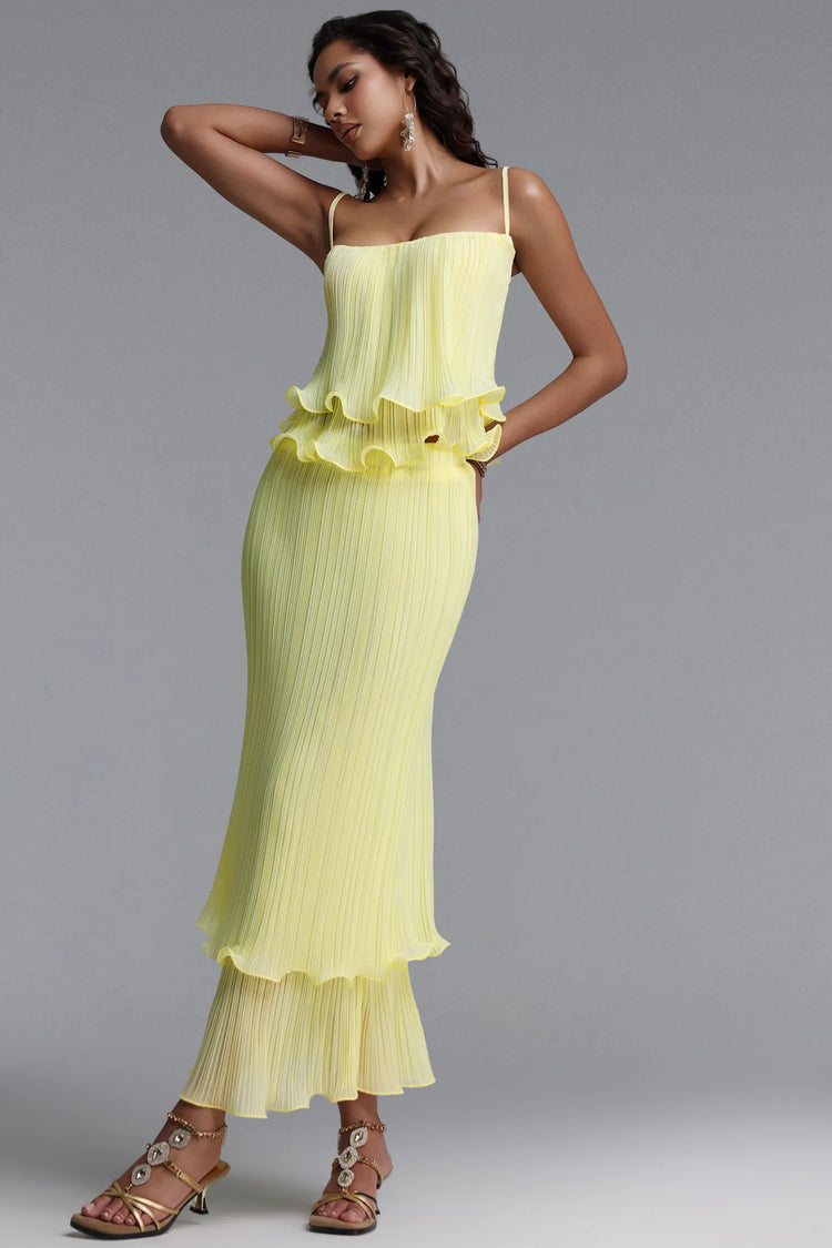 A woman modeling a pale yellow pleated midi dress with spaghetti straps, a ruffled peplum waist, and a tiered skirt, paired with large statement earrings and strappy sandals, against a solid gray studio background.