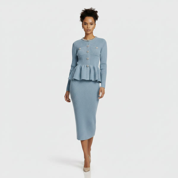 A model stands against a light gray studio background wearing a light blue ribbed knit two-piece set consisting of a long-sleeve cropped cardigan with a peplum hem and matching knee-length skirt.